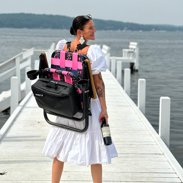 ParkIt Chair with the signature Nora fleming pattern with a cooler and drink holder nora is wearing it with the backpack straps on the dock at the lake