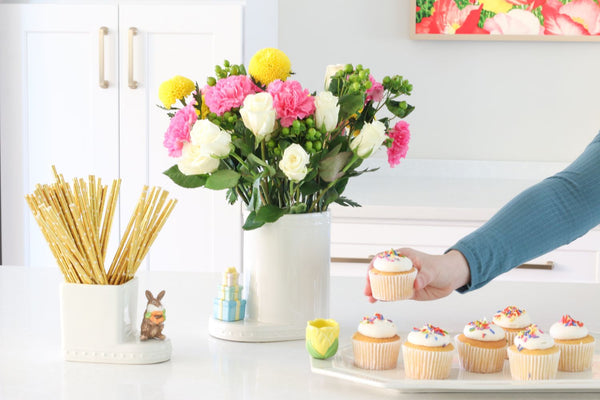 tulip mini on a octagonal platter with cupcakes, stack of presents on the utensil crock filled with flowers and bunny on a cutie container filled with straws