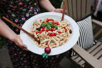 Person holding a plate of pasta salad with cherry tomatoes and basil leaves and a radish mini.