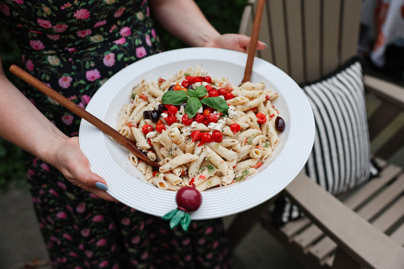 Person holding a plate of pasta salad with cherry tomatoes and basil leaves and a radish mini.