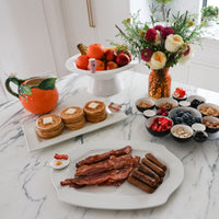 Breakfast spread with bacon, pancakes, and fruit on a marble table.
