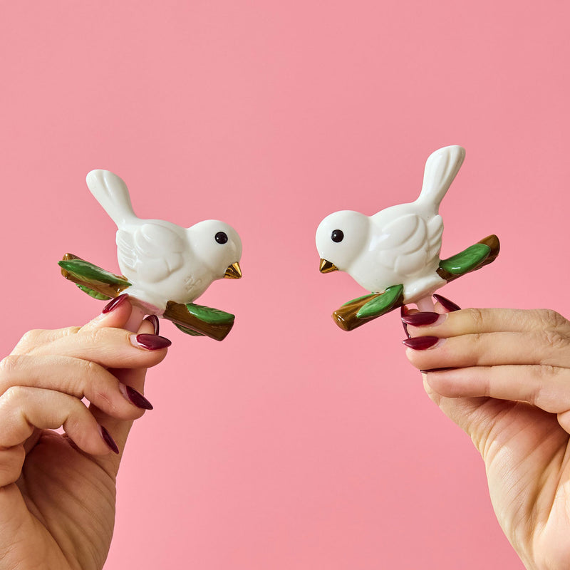White ceramic bird minsi on a branch with a pink background and hands holding them up