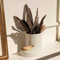 White utensil holder with feathers on a white surface next to a gold-framed mirror.