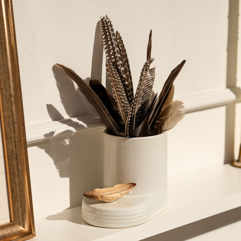 White utensil holder with feathers on a white surface next to a gold-framed mirror.