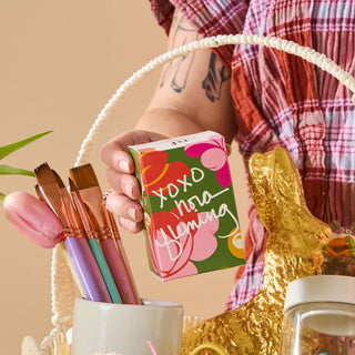 Person holding a box with floral design and text, surrounded by decorative items on a beige background.