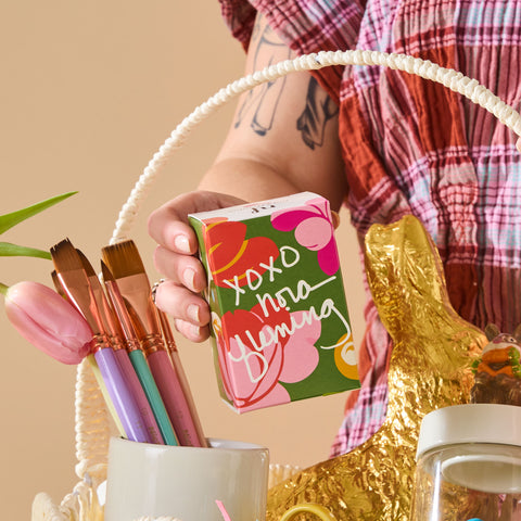 Person holding a box with floral design and text, surrounded by decorative items on a beige background.