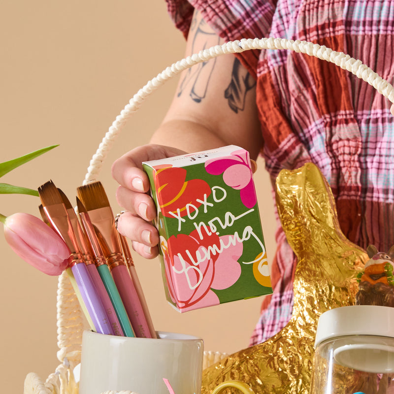 Person holding a box with floral design and text, surrounded by decorative items on a beige background.