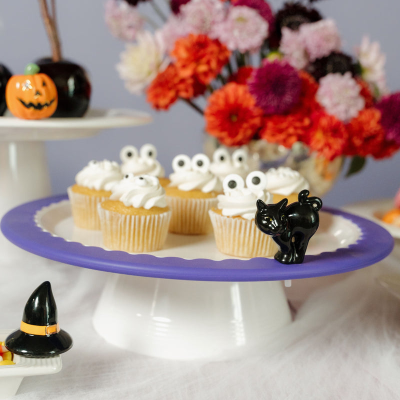 A black cat figurine with a glossy finish and yellow eyes, designed for decoration, possibly for Halloween. The mini sits on round platter on a low riser with cupcakes and floral arrangement in the background.