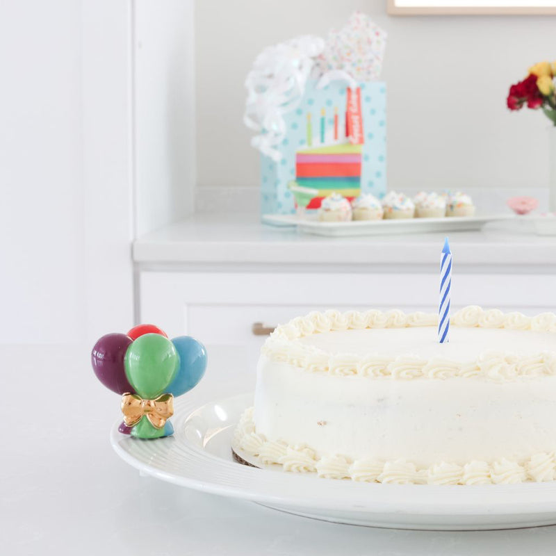 A ceramic mini four colorful balloons wrapped in  a gold bow on a round platter that holds a white birthday cake with one candle.
