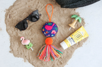 El Fuego Que Nos Unio round bird tassel on sand next to our flamingo mini, palm tree mini, sunscreen and sunglasses