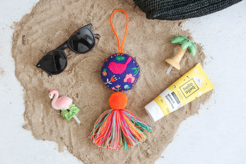 El Fuego Que Nos Unio round bird tassel on sand next to our flamingo mini, palm tree mini, sunscreen and sunglasses