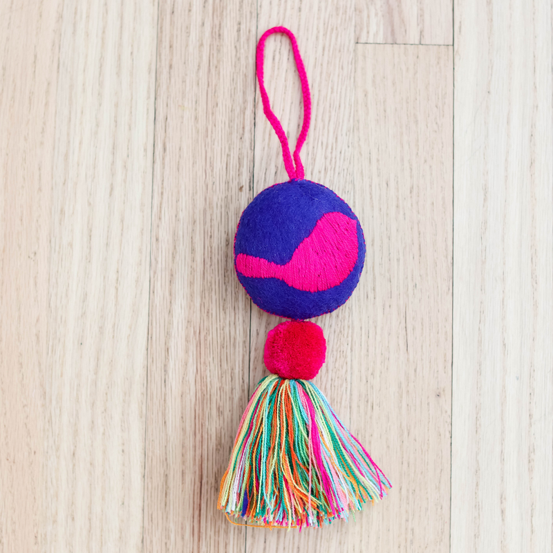 This image features a vibrant and colorful round bird tassel named "El Fuego Que Nos Unio." The tassel showcases a large, hand-embroidered circular design in bold blue and pink hues, topped with a bright pink pom-pom.